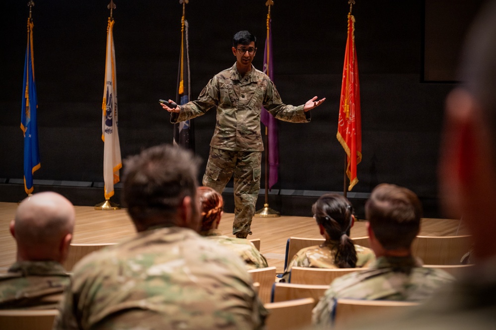 A military service member in OCP (Operational Camouflage Pattern) uniform is presenting or speaking to a seated audience of fellow service members in what appears to be a formal briefing room or auditorium. The speaker stands on a raised wooden stage with arms outstretched in an expressive gesture. Multiple military branch flags are displayed in the background.