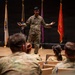 A military service member in OCP (Operational Camouflage Pattern) uniform is presenting or speaking to a seated audience of fellow service members in what appears to be a formal briefing room or auditorium. The speaker stands on a raised wooden stage with arms outstretched in an expressive gesture. Multiple military branch flags are displayed in the background.