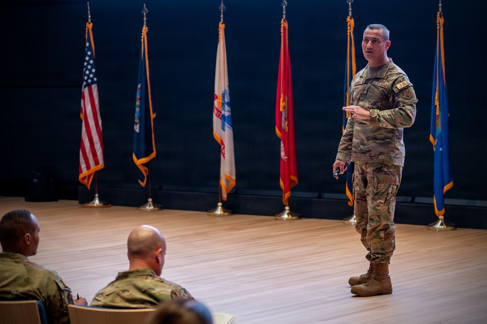 A military service member in OCP (Operational Camouflage Pattern) uniform is presenting or briefing on a stage to an audience of at least two other uniformed personnel. The setting appears to be a formal military auditorium or briefing hall with multiple service flags displayed in the background. The presenter holds what appears to be a small device, likely a presentation clicker.