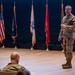 A military service member in OCP (Operational Camouflage Pattern) uniform is presenting or briefing on a stage to an audience of at least two other uniformed personnel. The setting appears to be a formal military auditorium or briefing hall with multiple service flags displayed in the background. The presenter holds what appears to be a small device, likely a presentation clicker.