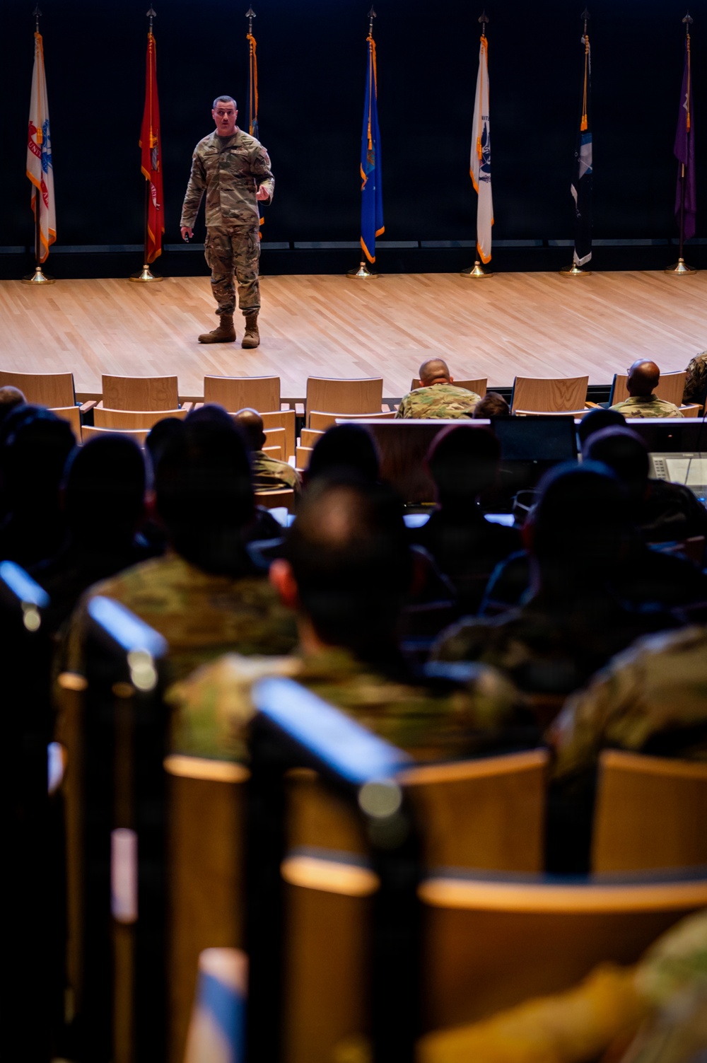 A military officer in OCP (Operational Camouflage Pattern) uniform stands on a stage addressing an audience of seated military personnel in what appears to be a formal auditorium or lecture hall. Multiple military unit flags and guidons are displayed on stands behind the speaker on the stage. The image is taken from the rear of the audience, providing a perspective shot looking toward the stage.