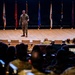 A military officer in OCP (Operational Camouflage Pattern) uniform stands on a stage addressing an audience of seated military personnel in what appears to be a formal auditorium or lecture hall. Multiple military unit flags and guidons are displayed on stands behind the speaker on the stage. The image is taken from the rear of the audience, providing a perspective shot looking toward the stage.