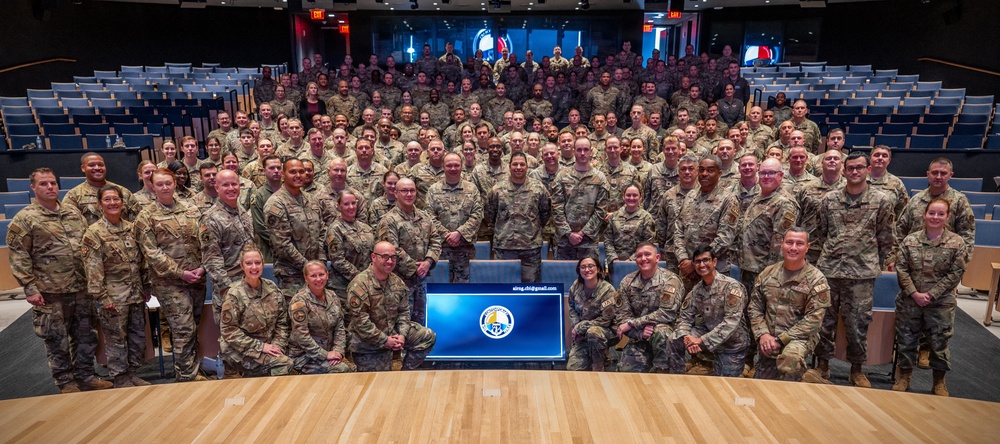 A large group photograph of military personnel, all wearing camouflage uniforms (OCP/MultiCam pattern), posed in a tiered auditorium or theater setting. A display screen in the foreground shows a military unit insignia and an email address. The group consists of approximately 150-200 service members arranged in multiple rows across the venue.