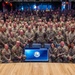 A large group photograph of military personnel, all wearing camouflage uniforms (OCP/MultiCam pattern), posed in a tiered auditorium or theater setting. A display screen in the foreground shows a military unit insignia and an email address. The group consists of approximately 150-200 service members arranged in multiple rows across the venue.