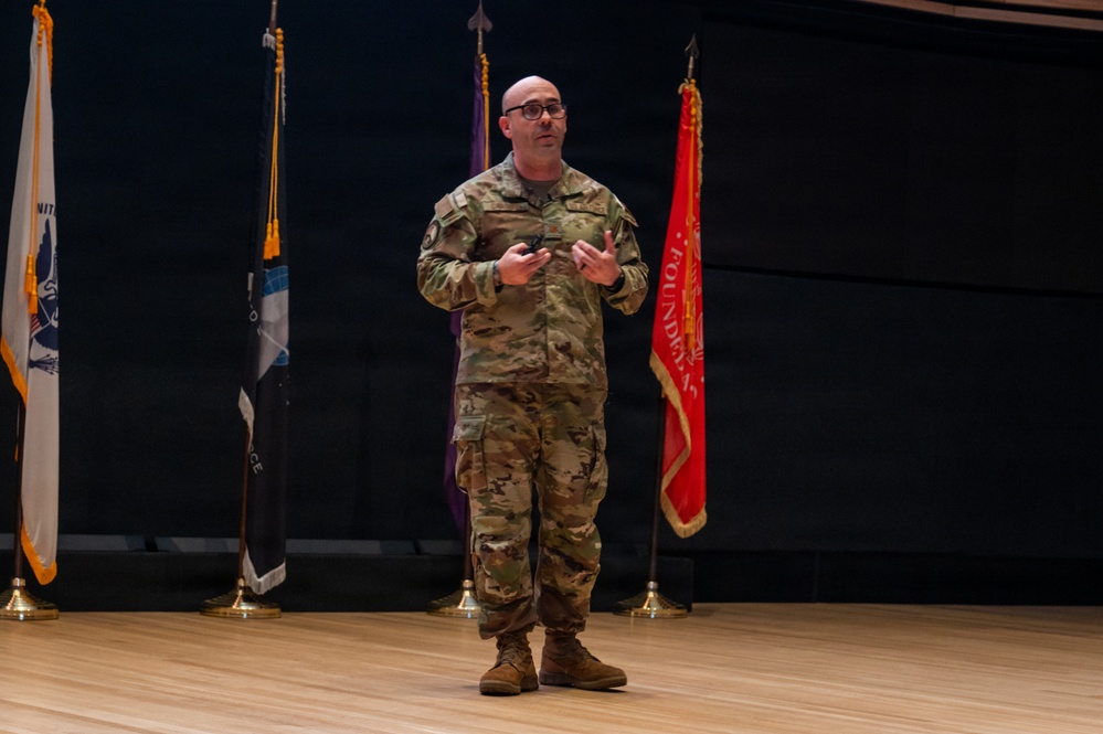 A bald male military personnel wearing OCP (Operational Camouflage Pattern) uniform is standing on a wooden stage, appearing to give a presentation or speech. He is holding what appears to be a presentation clicker in his hands and is gesturing while speaking. Several military and organizational flags are displayed on stands behind him against a dark backdrop.
