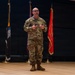 A bald male military personnel wearing OCP (Operational Camouflage Pattern) uniform is standing on a wooden stage, appearing to give a presentation or speech. He is holding what appears to be a presentation clicker in his hands and is gesturing while speaking. Several military and organizational flags are displayed on stands behind him against a dark backdrop.
