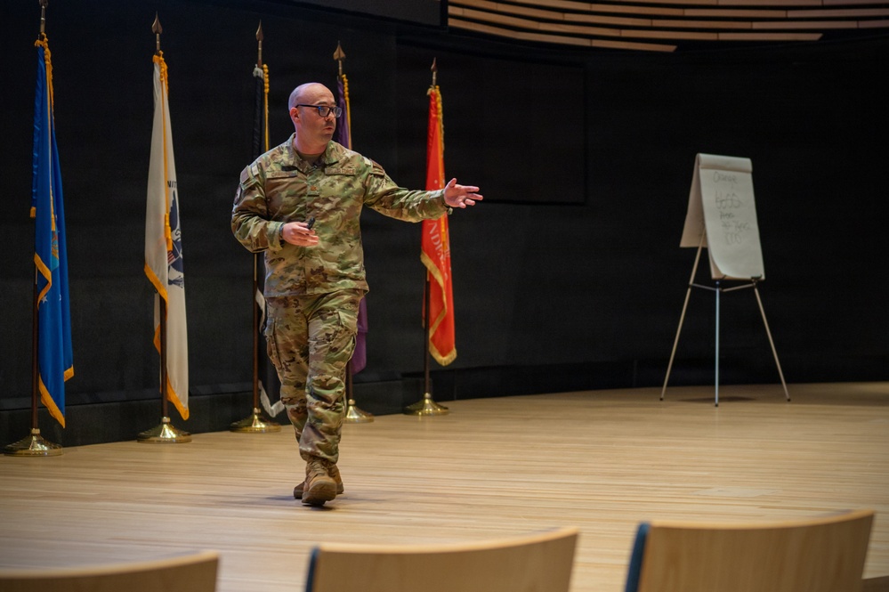A bald military officer wearing camouflage (OCP/MultiCam) uniform is presenting or briefing on a stage or auditorium floor. He is gesturing with his right arm outstretched, appearing to address an audience. A whiteboard easel with handwritten notes and multiple military flags on stands are visible in the background.