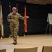 A bald military officer wearing camouflage (OCP/MultiCam) uniform is presenting or briefing on a stage or auditorium floor. He is gesturing with his right arm outstretched, appearing to address an audience. A whiteboard easel with handwritten notes and multiple military flags on stands are visible in the background.