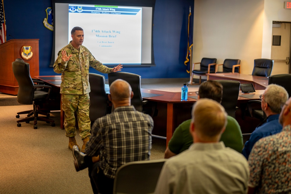 A military officer in OCP (Operational Camouflage Pattern) uniform is delivering a mission brief to a group of civilians in a conference room. The presentation slide behind him reads '174th Attack Wing Mission Brief' with a commander's name partially visible. The setting appears to be an official military briefing room with Air National Guard insignia visible.