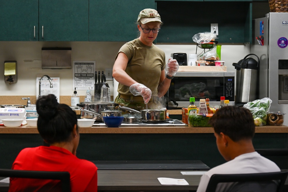 A woman in military-style clothing and a camouflage cap is conducting a cooking demonstration in a kitchen classroom setting. She is wearing disposable gloves and working with pots on a stovetop while two observers watch from the other side of a counter. The setting appears to be an instructional or community kitchen with various ingredients and condiments visible on the counter.