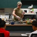 A woman in military-style clothing and a camouflage cap is conducting a cooking demonstration in a kitchen classroom setting. She is wearing disposable gloves and working with pots on a stovetop while two observers watch from the other side of a counter. The setting appears to be an instructional or community kitchen with various ingredients and condiments visible on the counter.