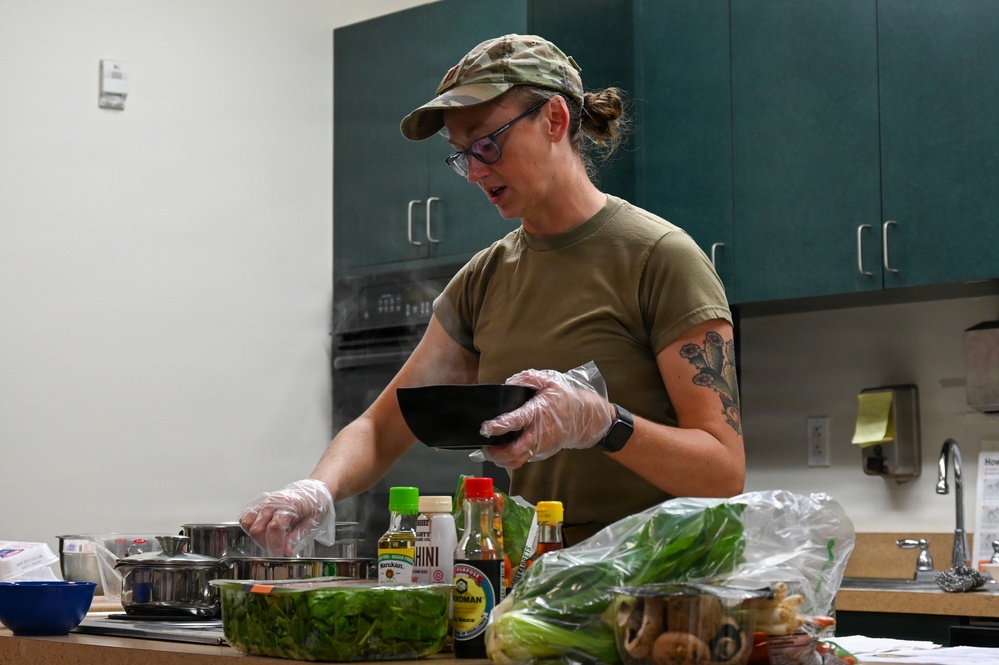 A woman wearing military-style clothing (olive green t-shirt and camouflage cap) is cooking in a kitchen while wearing disposable gloves. She is holding a black bowl and appears to be preparing a meal with various vegetables and condiments spread across the counter. A tattoo is visible on her right arm.
