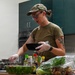 A woman wearing military-style clothing (olive green t-shirt and camouflage cap) is cooking in a kitchen while wearing disposable gloves. She is holding a black bowl and appears to be preparing a meal with various vegetables and condiments spread across the counter. A tattoo is visible on her right arm.