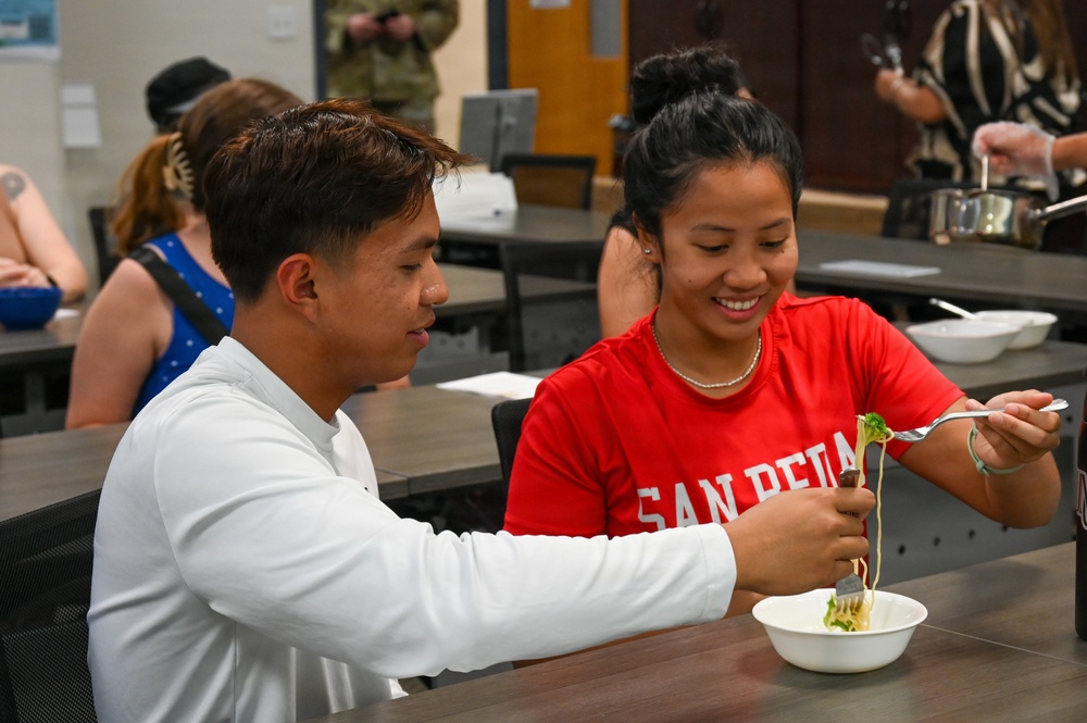 Two young people are seated at a table in what appears to be a cafeteria or dining hall, sharing a meal together. The woman in a red 'San Pedro' shirt is smiling and holding a fork with food, while the man in a white long-sleeve shirt is using a fork in a shared bowl. The setting suggests a casual communal dining environment with other people visible in the background.