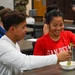 Two young people are seated at a table in what appears to be a cafeteria or dining hall, sharing a meal together. The woman in a red 'San Pedro' shirt is smiling and holding a fork with food, while the man in a white long-sleeve shirt is using a fork in a shared bowl. The setting suggests a casual communal dining environment with other people visible in the background.