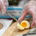 A person wearing disposable plastic gloves is cutting a hard-boiled egg on a wooden cutting board. The egg has been sliced to reveal a fully cooked yolk with a slightly jammy center. A box of disposable gloves is visible in the background, suggesting a food service or food safety context.