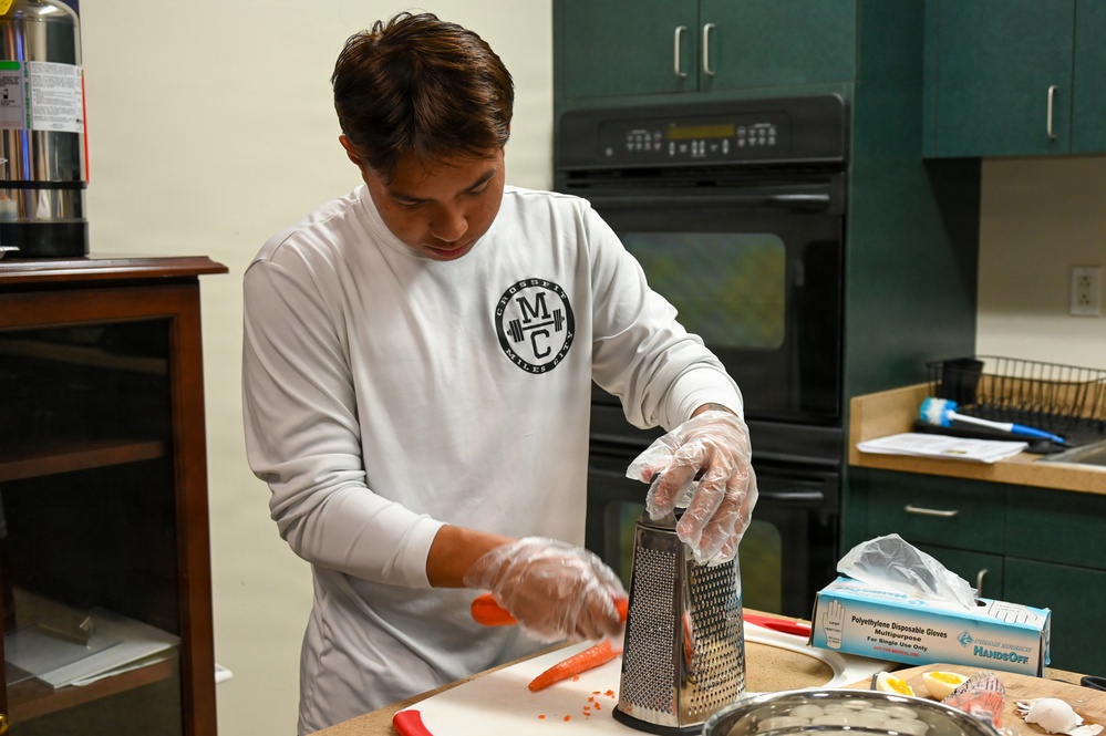 A young man wearing a white long-sleeve CrossFit shirt and disposable plastic gloves is grating carrots on a box grater at a kitchen counter. The kitchen features dark green cabinetry and a black double wall oven in the background. Several food preparation items and a box of disposable gloves are visible on the counter.