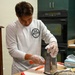 A young man wearing a white long-sleeve CrossFit shirt and disposable plastic gloves is grating carrots on a box grater at a kitchen counter. The kitchen features dark green cabinetry and a black double wall oven in the background. Several food preparation items and a box of disposable gloves are visible on the counter.