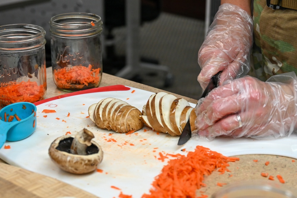 A person wearing disposable plastic gloves and a camouflage uniform is slicing a potato in a hasselback or accordion style on a white cutting board. The workspace also contains shredded carrots, a portobello mushroom cap, Mason jars filled with shredded carrots, and a blue measuring cup. The setting appears to be a food preparation area, likely a military or institutional kitchen.