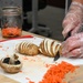 A person wearing disposable plastic gloves and a camouflage uniform is slicing a potato in a hasselback or accordion style on a white cutting board. The workspace also contains shredded carrots, a portobello mushroom cap, Mason jars filled with shredded carrots, and a blue measuring cup. The setting appears to be a food preparation area, likely a military or institutional kitchen.