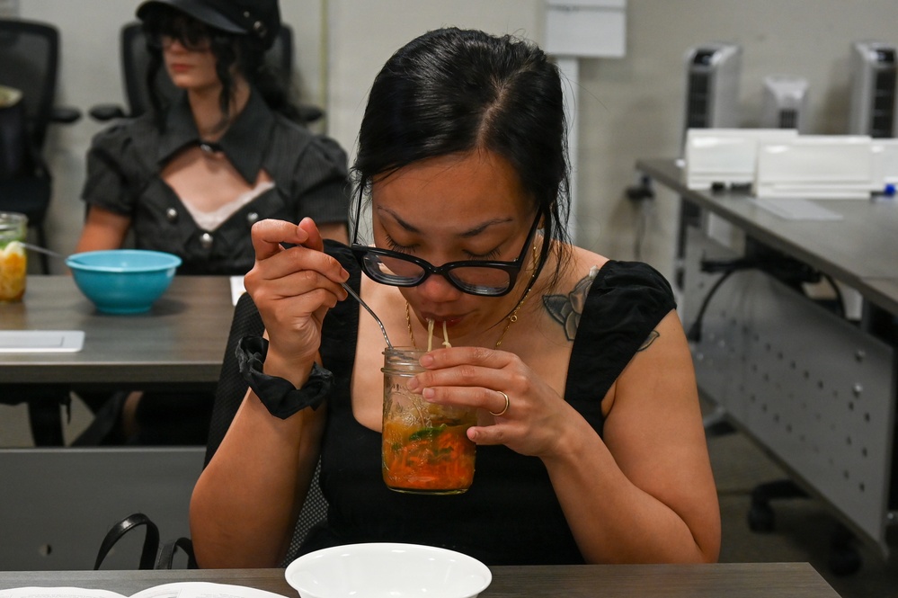A woman with black hair, glasses, and visible tattoos on her shoulder is leaning over a mason jar containing an orange-colored food or drink, using a spoon and straw simultaneously. She is seated at a table in what appears to be a classroom or office setting. In the background, another person dressed in all black with a wide-brimmed hat is seated at a separate table with a teal bowl.