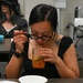 A woman with black hair, glasses, and visible tattoos on her shoulder is leaning over a mason jar containing an orange-colored food or drink, using a spoon and straw simultaneously. She is seated at a table in what appears to be a classroom or office setting. In the background, another person dressed in all black with a wide-brimmed hat is seated at a separate table with a teal bowl.