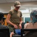 A person in military uniform (OCP/multicam pattern) stands at a table interacting with two seated civilians in what appears to be an indoor facility or community center. The uniformed individual is leaning over a blue bowl on the table, appearing to assist or demonstrate something. The setting suggests a community outreach, assistance, or educational event.