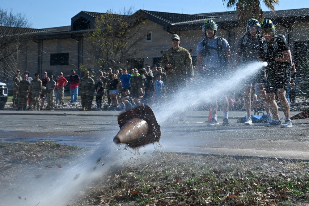 A water-filled football or similar object is bursting apart mid-air, spraying water in multiple directions near a curb. A crowd of people, many appearing to be in military or athletic attire, watches the event from the background. Two individuals in the foreground wear helmets and appear to be participants in the activity.