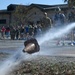 A water-filled football or similar object is bursting apart mid-air, spraying water in multiple directions near a curb. A crowd of people, many appearing to be in military or athletic attire, watches the event from the background. Two individuals in the foreground wear helmets and appear to be participants in the activity.