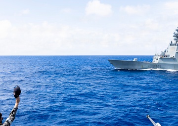 Naval personnel aboard one warship are waving farewell to a passing warship identified as F101, which is flying a Spanish flag. The scene depicts a maritime greeting or departure ceremony between two naval vessels at sea. A deck-mounted gun mount is visible in the foreground of the observing vessel.