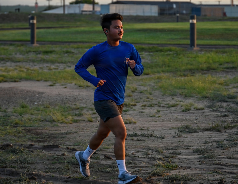 A young man is running outdoors on a dirt and grass path, wearing a blue long-sleeve athletic shirt, dark shorts, white socks, and gray running shoes. He has wireless earbuds in and appears to be mid-stride during a training run or workout. The setting appears to be an open field or athletic grounds during golden hour lighting.