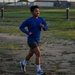 A young man is running outdoors on a dirt and grass path, wearing a blue long-sleeve athletic shirt, dark shorts, white socks, and gray running shoes. He has wireless earbuds in and appears to be mid-stride during a training run or workout. The setting appears to be an open field or athletic grounds during golden hour lighting.