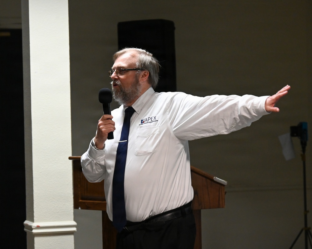 A middle-aged man with gray hair and a beard is speaking at a podium while holding a microphone. He is wearing a white dress shirt with an 'APEX' logo on the chest pocket and a navy blue tie. His right arm is extended outward in an expressive gesture, suggesting an animated public presentation or speech.