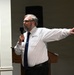 A middle-aged man with gray hair and a beard is speaking at a podium while holding a microphone. He is wearing a white dress shirt with an 'APEX' logo on the chest pocket and a navy blue tie. His right arm is extended outward in an expressive gesture, suggesting an animated public presentation or speech.