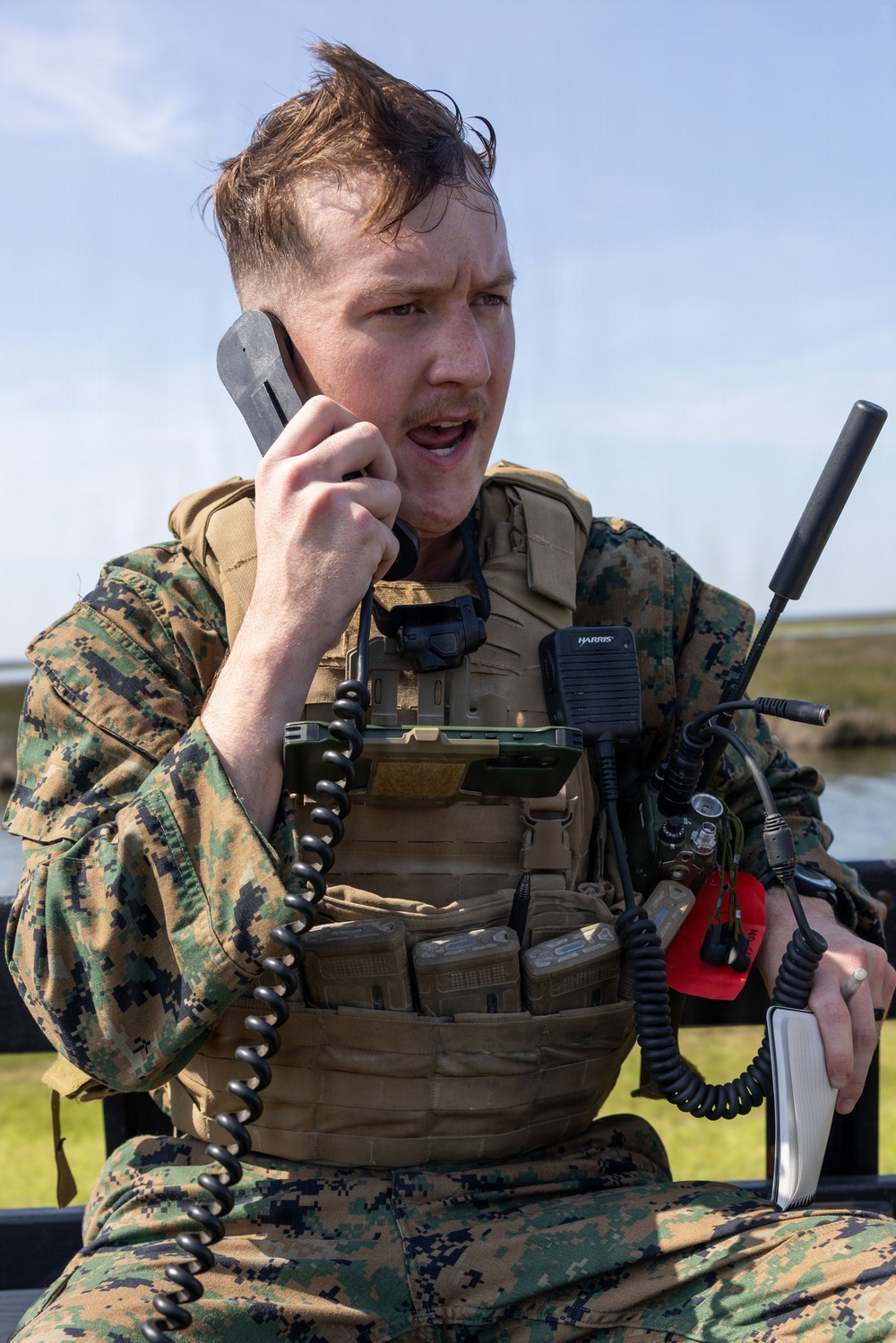A male military personnel wearing MARPAT (Marine pattern) digital camouflage uniform and tactical plate carrier vest is actively communicating via a corded handset. He is equipped with multiple communication devices including a Harris radio with antenna and additional handheld equipment. The outdoor setting appears to be a coastal or wetland environment under clear skies.