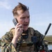 A male military personnel wearing MARPAT (Marine pattern) digital camouflage uniform and tactical plate carrier vest is actively communicating via a corded handset. He is equipped with multiple communication devices including a Harris radio with antenna and additional handheld equipment. The outdoor setting appears to be a coastal or wetland environment under clear skies.