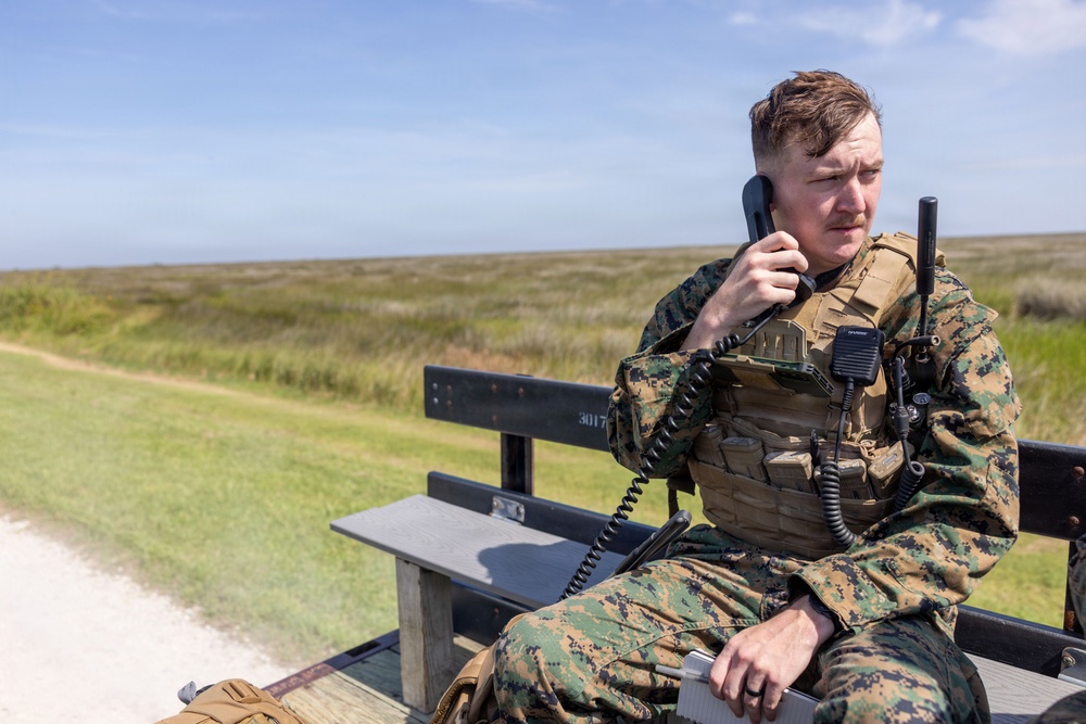A U.S. Marine in MARPAT woodland digital camouflage uniform and tactical plate carrier vest is seated on a wooden bench, using a corded radio handset. He appears to be actively communicating, with additional radio equipment mounted on his vest. The setting is an open grassy field with a dirt path visible in the background under a clear blue sky.