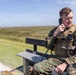 A U.S. Marine in MARPAT woodland digital camouflage uniform and tactical plate carrier vest is seated on a wooden bench, using a corded radio handset. He appears to be actively communicating, with additional radio equipment mounted on his vest. The setting is an open grassy field with a dirt path visible in the background under a clear blue sky.