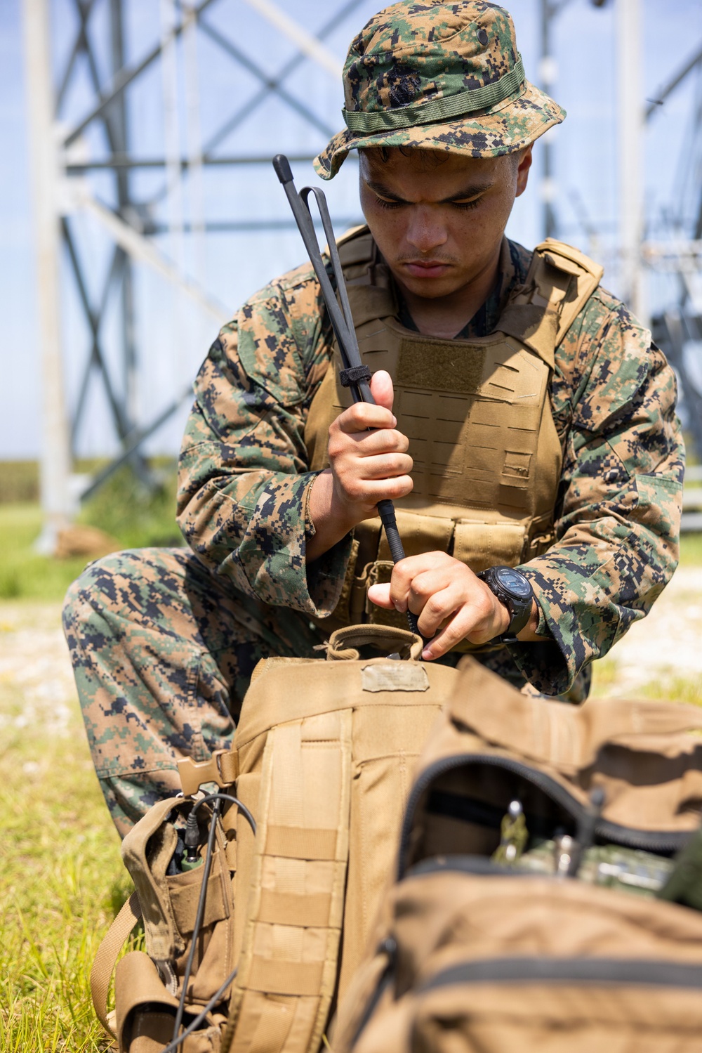 A U.S. Marine in MARPAT woodland digital camouflage uniform is seated outdoors, handling what appears to be a radio antenna or communications device over a tan tactical pack. He is wearing a plate carrier vest, boonie cover, and a tactical watch, suggesting a field communications or training exercise. A large antenna tower or transmission structure is visible in the background.