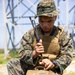 A U.S. Marine in MARPAT woodland digital camouflage uniform is seated outdoors, handling what appears to be a radio antenna or communications device over a tan tactical pack. He is wearing a plate carrier vest, boonie cover, and a tactical watch, suggesting a field communications or training exercise. A large antenna tower or transmission structure is visible in the background.