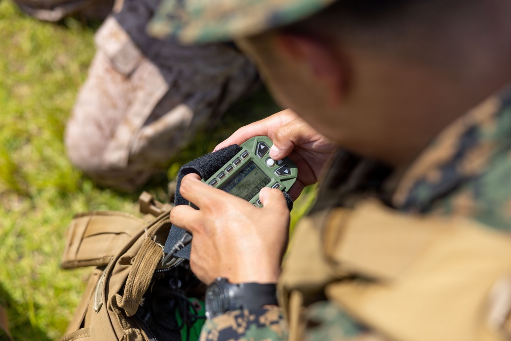 A military personnel in digital camouflage uniform is operating a small green tactical electronic device, likely a GPS unit or radio controller. The soldier is seated outdoors on grass, with a tan/coyote tactical backpack visible nearby. The image is captured from an over-the-shoulder perspective with shallow depth of field.