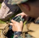 A military personnel in digital camouflage uniform is operating a small green tactical electronic device, likely a GPS unit or radio controller. The soldier is seated outdoors on grass, with a tan/coyote tactical backpack visible nearby. The image is captured from an over-the-shoulder perspective with shallow depth of field.