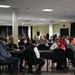 A large indoor meeting or briefing is taking place in what appears to be a conference or event hall. Attendees are seated at black-draped tables arranged in rows, facing toward a presenter or screen outside the frame. The audience includes a mix of civilians and military personnel in uniform.