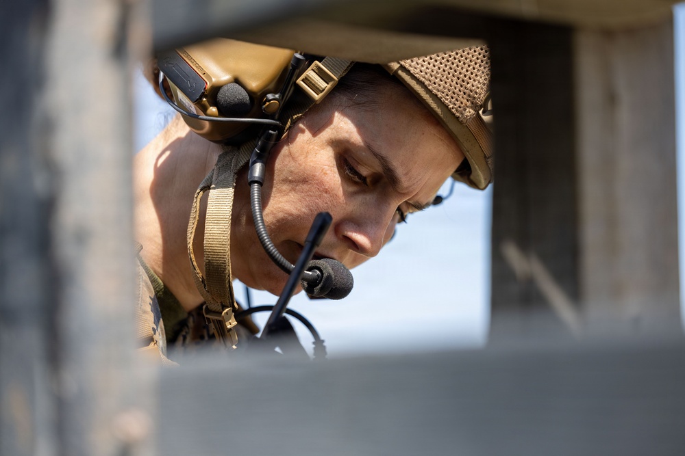 A military or tactical personnel is photographed in close-up through a rectangular opening or aperture in a concrete or stone structure. The individual is wearing tactical communications headgear with a boom microphone and appears to be focused downward, possibly monitoring equipment or a display. The framing through the structural opening creates a confined, operational context.