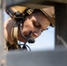 A military or tactical personnel is photographed in close-up through a rectangular opening or aperture in a concrete or stone structure. The individual is wearing tactical communications headgear with a boom microphone and appears to be focused downward, possibly monitoring equipment or a display. The framing through the structural opening creates a confined, operational context.