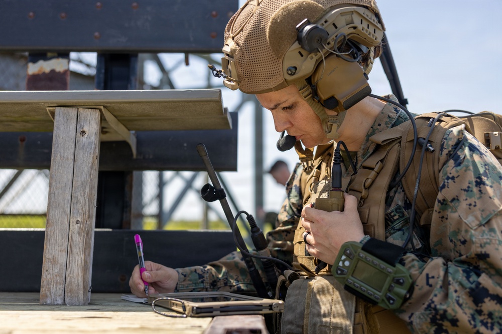 A military service member in full combat gear is seated at a wooden table, writing notes with a pink pen while holding a radio handset. The individual is wearing MARPAT (Marine pattern) camouflage uniform, a tan tactical helmet with communications headset, and is equipped with multiple radios and tactical gear. The setting appears to be an outdoor training or operational environment.