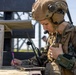 A military service member in full combat gear is seated at a wooden table, writing notes with a pink pen while holding a radio handset. The individual is wearing MARPAT (Marine pattern) camouflage uniform, a tan tactical helmet with communications headset, and is equipped with multiple radios and tactical gear. The setting appears to be an outdoor training or operational environment.