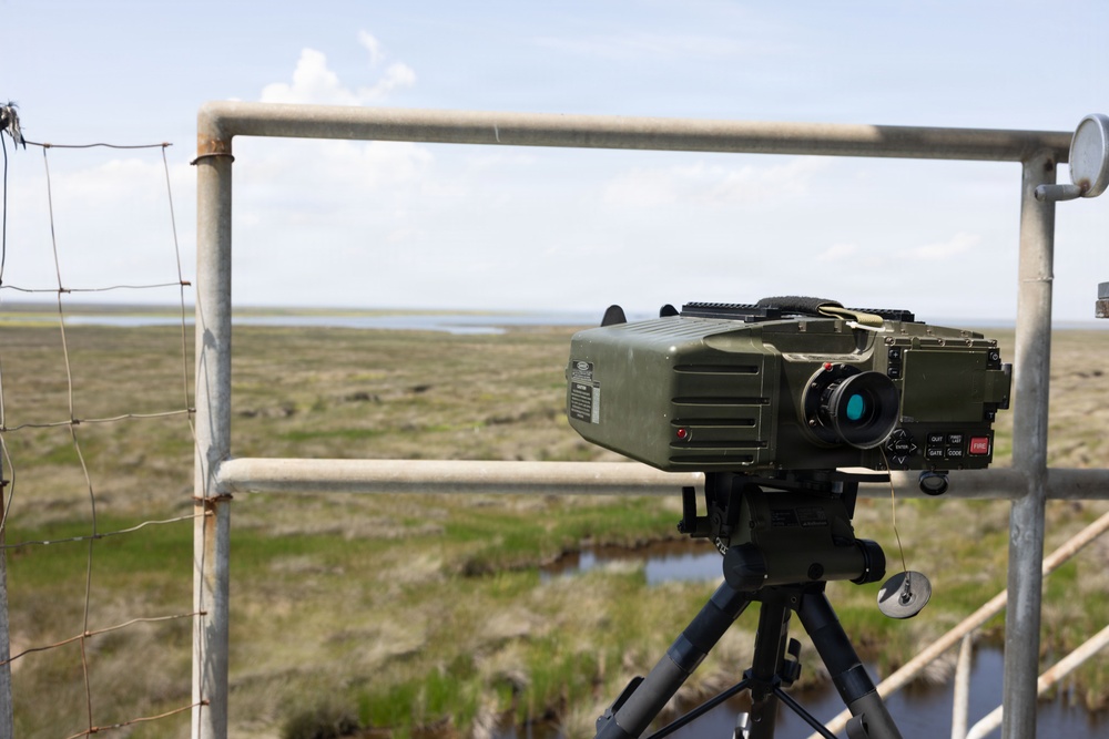 A military-grade optical or thermal imaging device mounted on a black tripod is positioned on an elevated platform overlooking a flat, marshy coastal landscape. The device is olive/military green in color and features a prominent teal-coated lens, control buttons, and indicator lights. The setting appears to be a surveillance or observation post in a wetland or estuary environment.