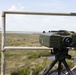 A military-grade optical or thermal imaging device mounted on a black tripod is positioned on an elevated platform overlooking a flat, marshy coastal landscape. The device is olive/military green in color and features a prominent teal-coated lens, control buttons, and indicator lights. The setting appears to be a surveillance or observation post in a wetland or estuary environment.