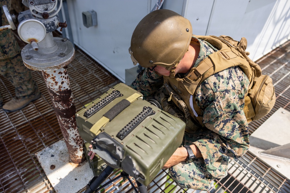 A U.S. Marine in full combat gear is closely inspecting or operating a military equipment case or device on what appears to be the deck of a naval vessel or industrial platform. The Marine is wearing MARPAT digital camouflage, a Kevlar helmet, and a tactical vest with a backpack. A second Marine is partially visible in the background.